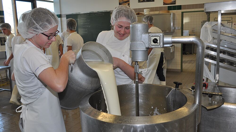 Zwei Schüler gießen Milch von einer Milchkanne in einen großen Kessel