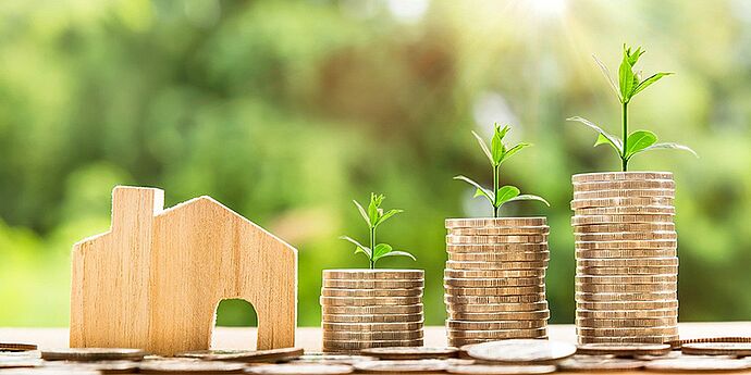 (cc) pixabay.com/nattan23 Ein kleines Haus aus Holz. Rechts daneben drei höher werdene Münztürme aus welchen jeweils ein Pflänzchen sprießt.