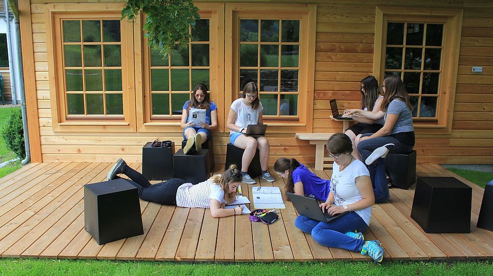 Schülerinnen sitzen auf Holzterrasse und arbeiten am Laptop 