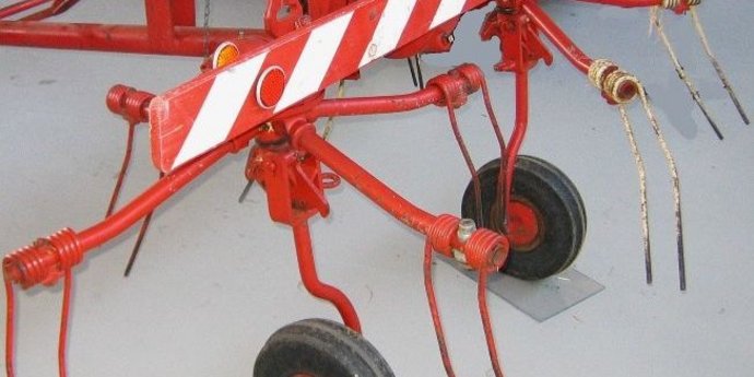 Rote landwirtschaftliche Maschine vom Typ Kreiselzettwender mit mehreren Zinkenrädern und kleinen Rädern steht auf hellem Boden.