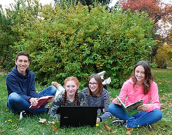Schülerinnen und Schüler sitzen mit Computer und Büchern im Park