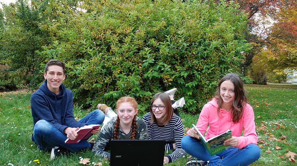 Schülerinnen und Schüler sitzen mit Computer und Büchern im Park
