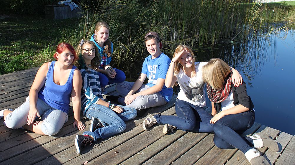 Schülerinnen und Schüler sitzen auf Steg am Teich