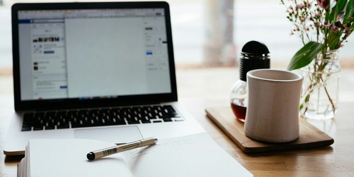 Laptop, Notizbuch und Kaffeetasse auf einem Schreibtisch vor einem Fenster – ein Arbeitsplatz zum Schreiben oder Arbeiten.