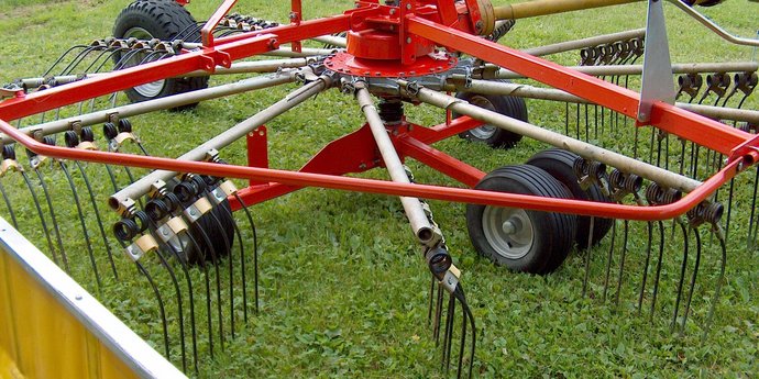 Eine große landwirtschaftliche Maschine mit roten Metallarmen und vielen schwarzen Zinken steht auf einer Wiese.